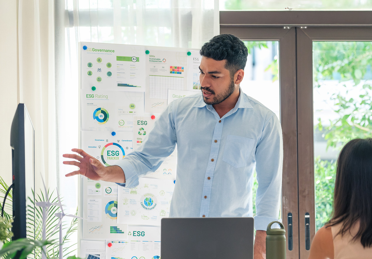 Homem apresentando gráficos sobre ESG e sustentabilidade em uma reunião, com notebook, monitor e quadro de dados.
