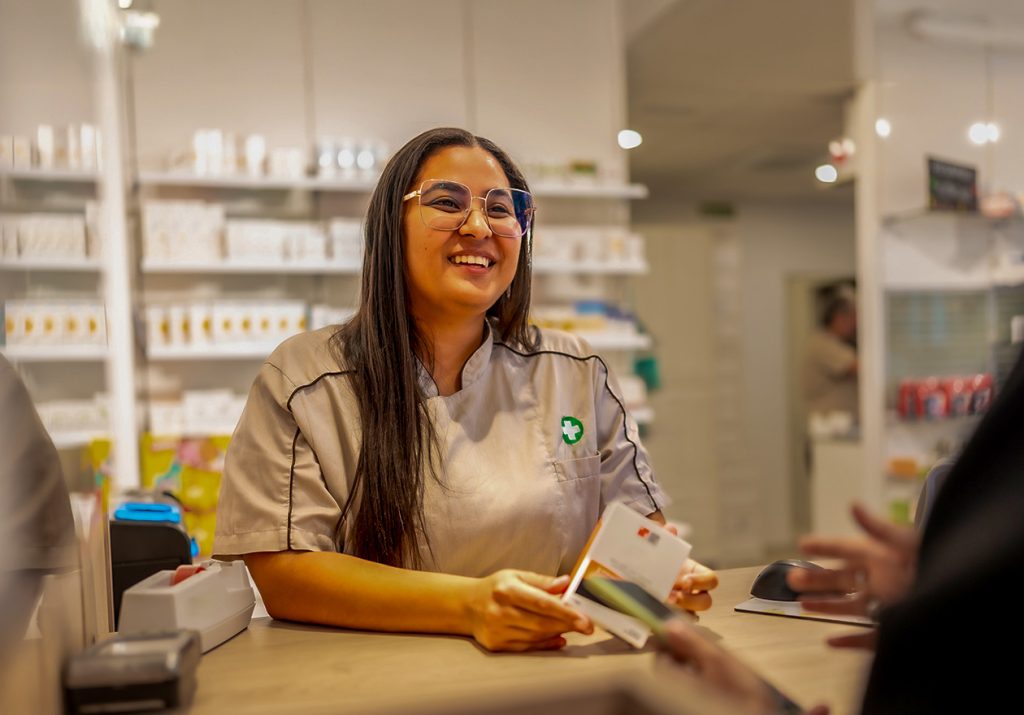 Uma farmacêutica sorridente entrega uma caixa de medicamento a um cliente em uma farmácia com prateleiras ao fundo. Curso: Farmácia Clínica.