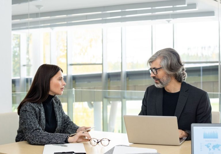 Duas pessoas conversam em um escritório com laptop, papéis e óculos sobre a mesa. Curso: Feedback Assertivo.