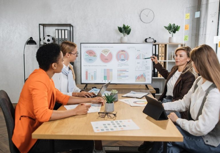 Um grupo de mulheres discute em uma reunião com gráficos na tela e papéis na mesa. Curso: Ferramentas de Gestão da Qualidade.