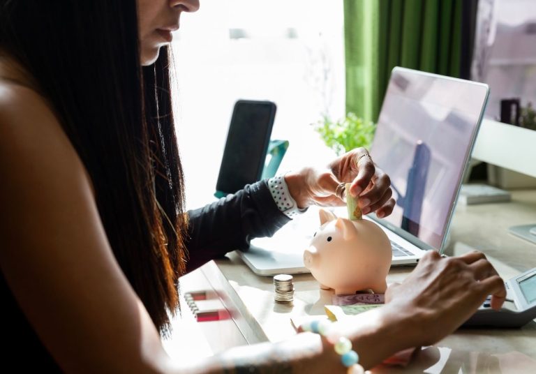 Uma mulher coloca moedas em um cofrinho de porquinho enquanto trabalha no laptop em uma mesa. Curso: Finanças Pessoais - Noções Básicas.