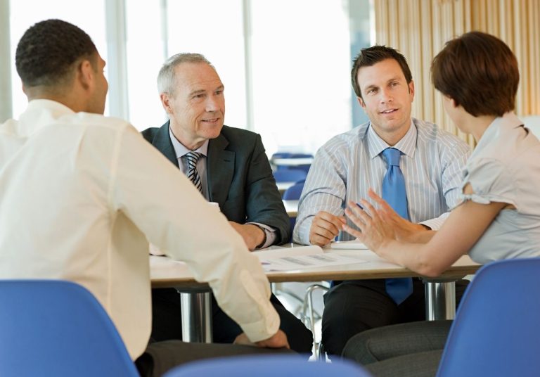 Quatro pessoas conversam em uma reunião, sentadas em cadeiras azuis, em uma sala ampla. Curso: Formação de Cooperados.