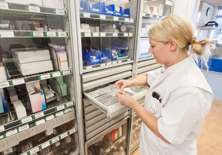Uma farmacêutica de jaleco organiza medicamentos em uma gaveta de farmácia hospitalar com prateleiras ao fundo. Curso: Fundamentos de Farmácia Hospitalar e Clínica.