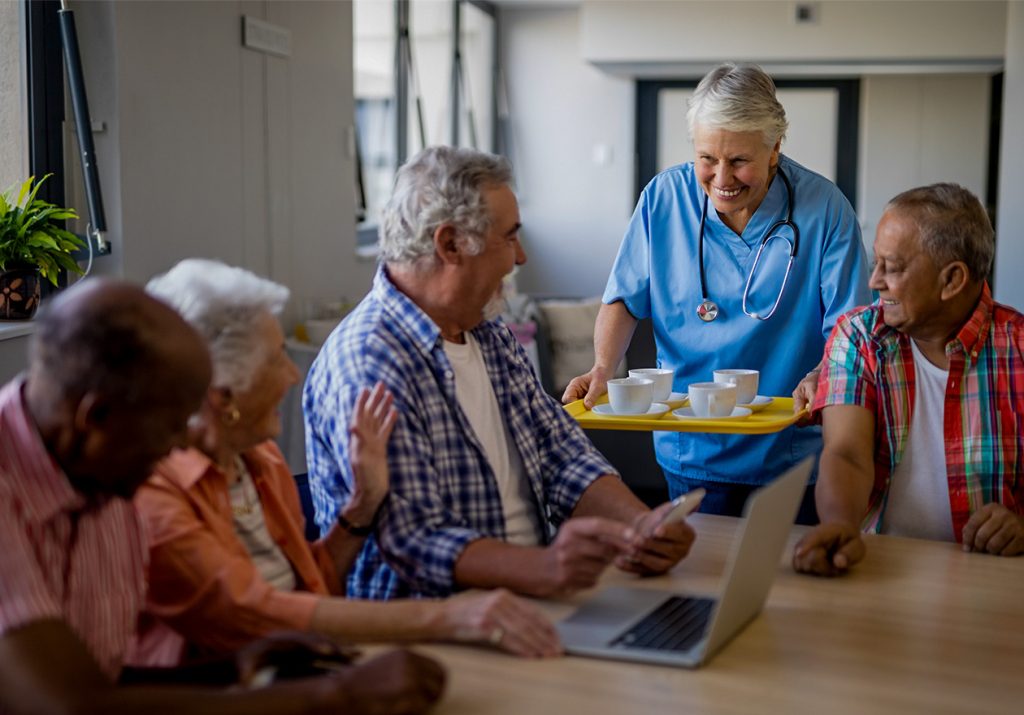 Uma enfermeira sorridente oferece xícaras a idosos reunidos em torno de uma mesa com laptop. Curso: Geriatria e Gerontologia para Entrega de Valor.