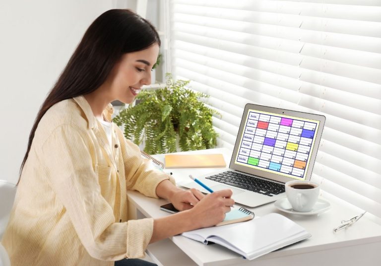 Mulher sorridente faz anotações em caderno, com laptop exibindo calendário, café e planta sobre a mesa. Curso: Gestão de Execução.