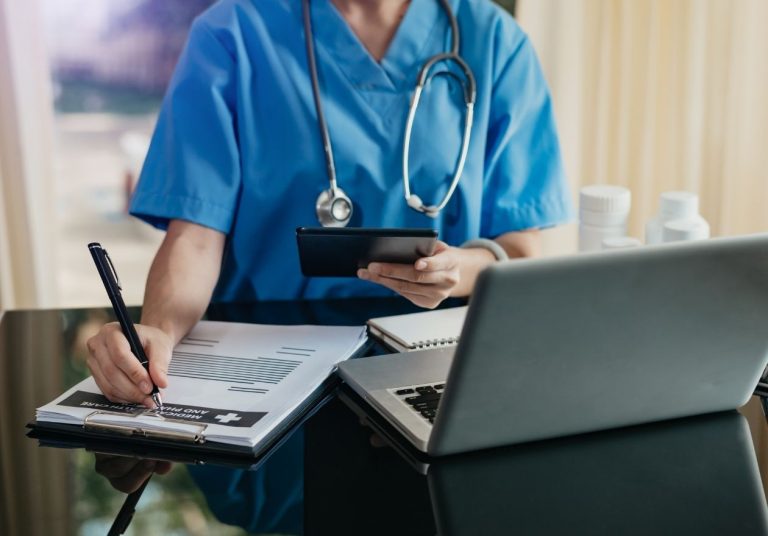 Profissional de saúde em uniforme azul escreve em documentos, com tablet na mão e estetoscópio sobre o pescoço. Curso: Introdução à Regulação dos Planos de Saúde.