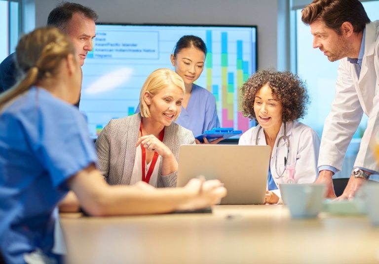 Equipe de saúde conversa ao redor de uma mesa com laptop e gráficos na tela ao fundo. Curso: Lean Health - Gestão de Fluxo Hospitalar.