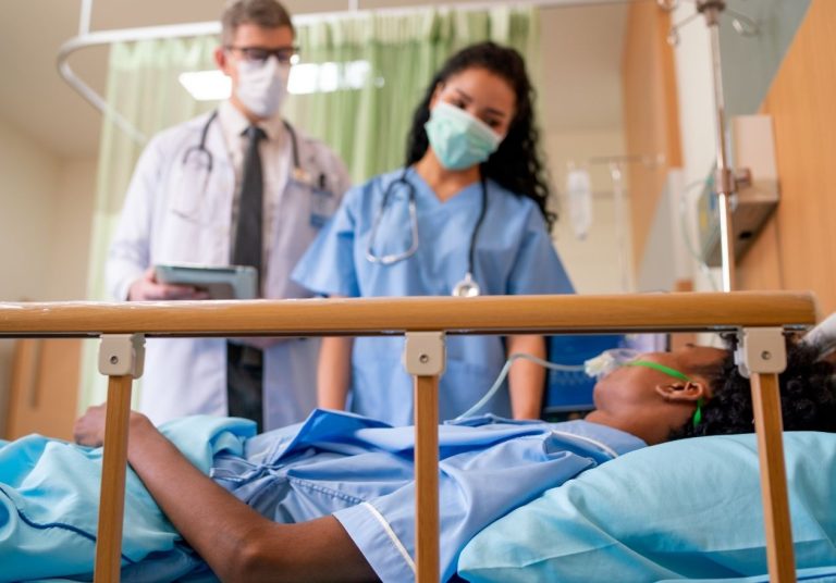 Profissionais de saúde atendem um paciente internado com suporte respiratório em uma cama hospitalar. Curso: Manejo Paciente Covid-19 (Avançado).