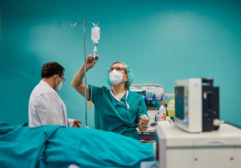 Profissionais de saúde ajustam um soro para um paciente em uma cama hospitalar, com equipamentos médicos ao lado. Curso: Manejo Paciente Covid-19 Básico.