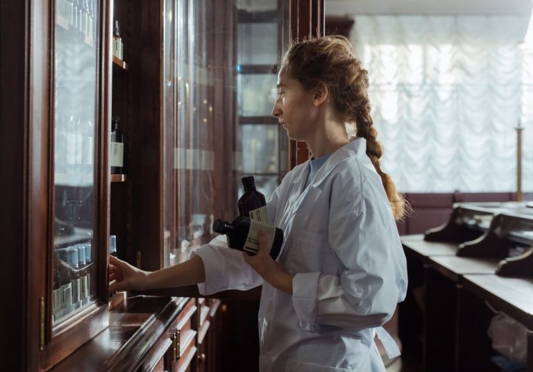 Farmacêutica manipula medicamentos, com frascos e equipamentos em um armário de madeira. Curso: Manipulação e Atenção Farmacêutica.