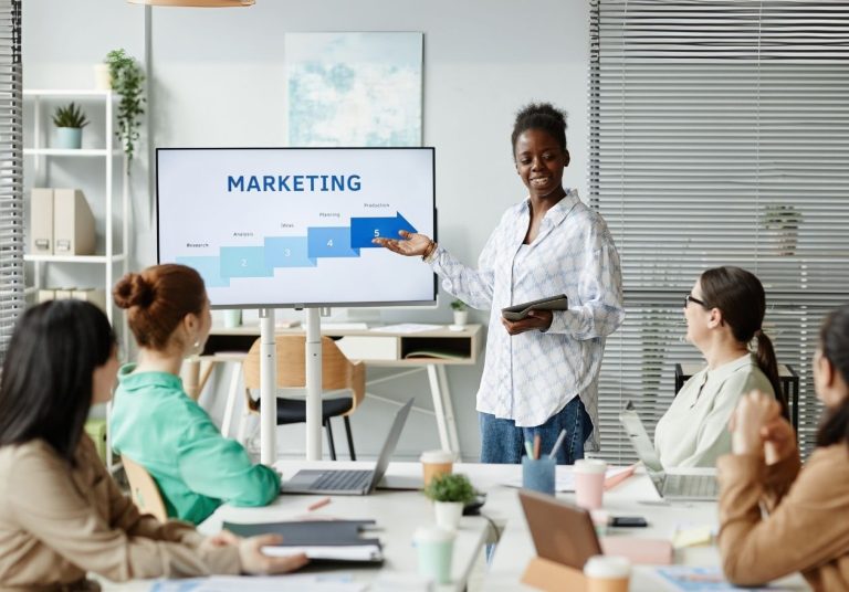 Mulher apresenta gráfico de marketing em uma tela para um grupo em sala de reunião. Curso: Marketing Digital.