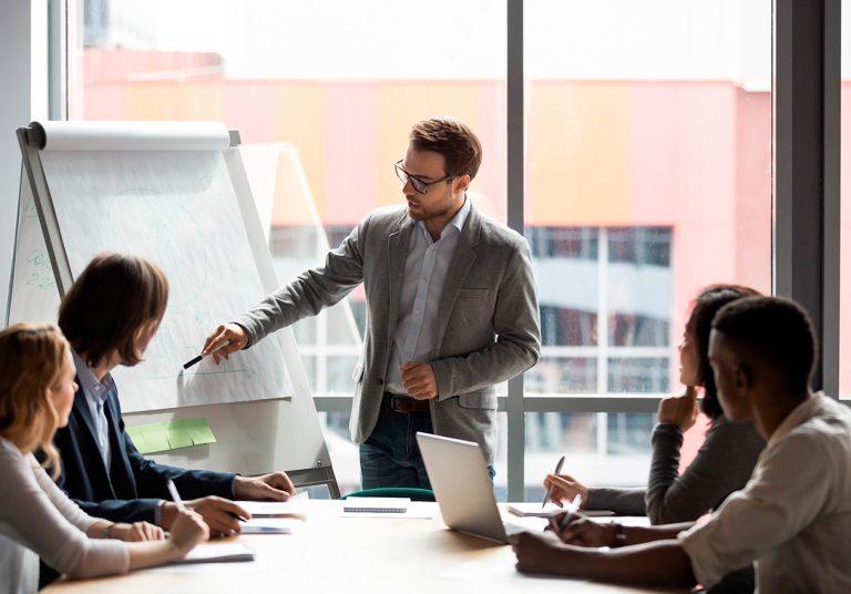 Equipe em reunião de trabalho sobre MBA em gestão de cooperativas, um homem apresenta informações em flip chart enquanto outros quatro, entre homens e mulheres, tomam notas.