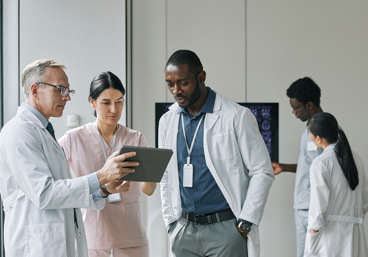 Equipe médica com três pessoas discutindo sobre o MBA em gestão hospitalar e organizações de saúde. Um dos homens segura um tablet onde mostra informações. No fundo, duas pessoas observando um monitor.