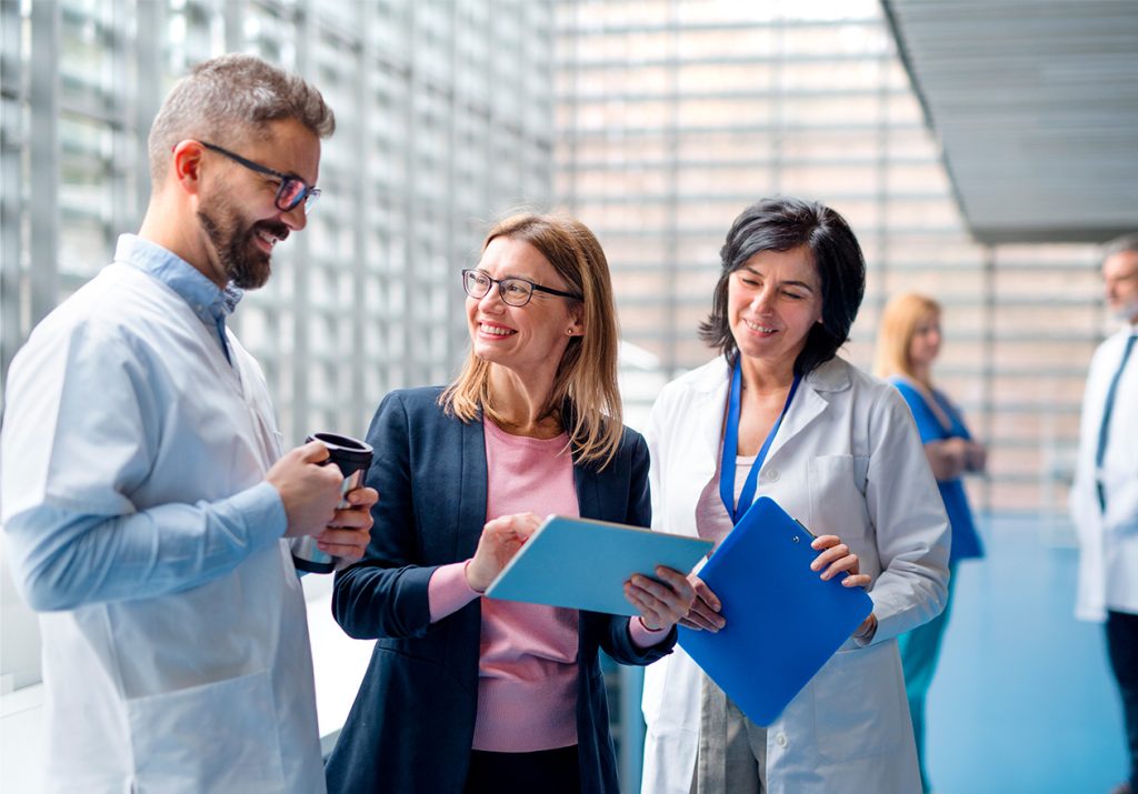 Profissionais da saúde conversando sobre o MBA em governança, riscos, regulação e compliance no corredor de um hospital, segurando prontuários e café em um ambiente moderno.