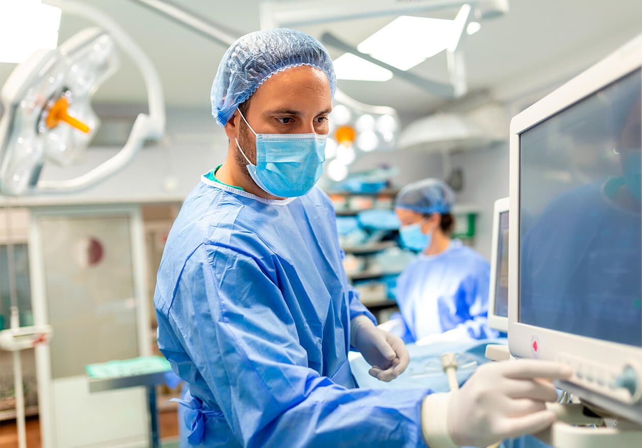 Sala de cirurgia com monitores, equipamentos médicos e iluminação cirúrgica. Há dois profissionais de medicina intensiva, no fundo uma mulher e na frente um homem. Ambos vestem roupas cirúrgicas azuis, máscara, touca e luvas.