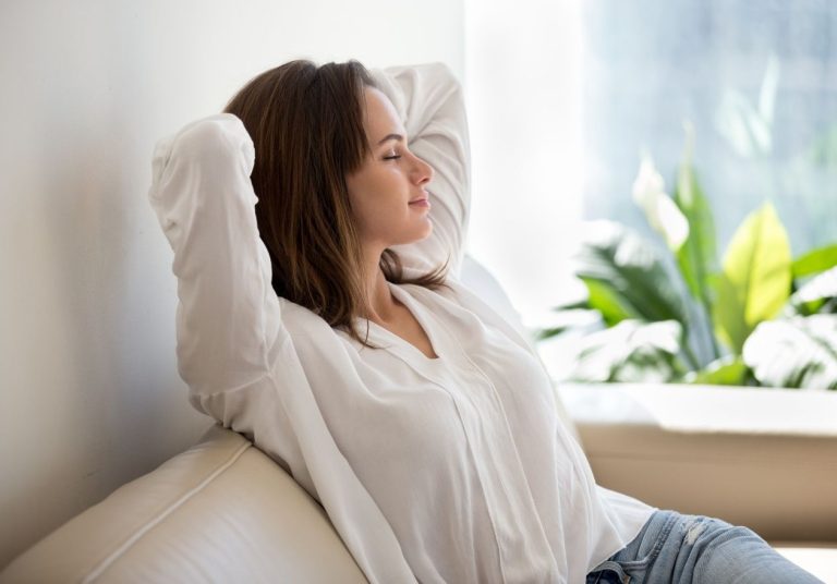 Uma mulher com os braços atrás da cabeça relaxa em um sofá, perto de uma planta. Curso: Mindfulness.