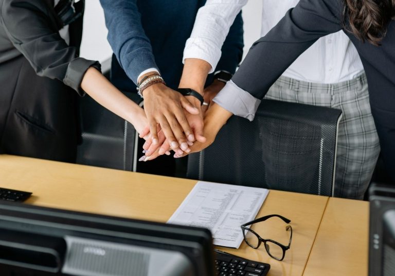 Quatro pessoas unem as mãos sobre uma mesa no escritório. Curso: Missão, Visão e Valores.