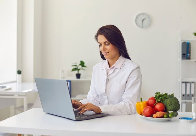 Uma profissional de jaleco trabalha em um laptop, com frutas e vegetais coloridos ao lado. Curso: Nutrição de Produção e Clínica.