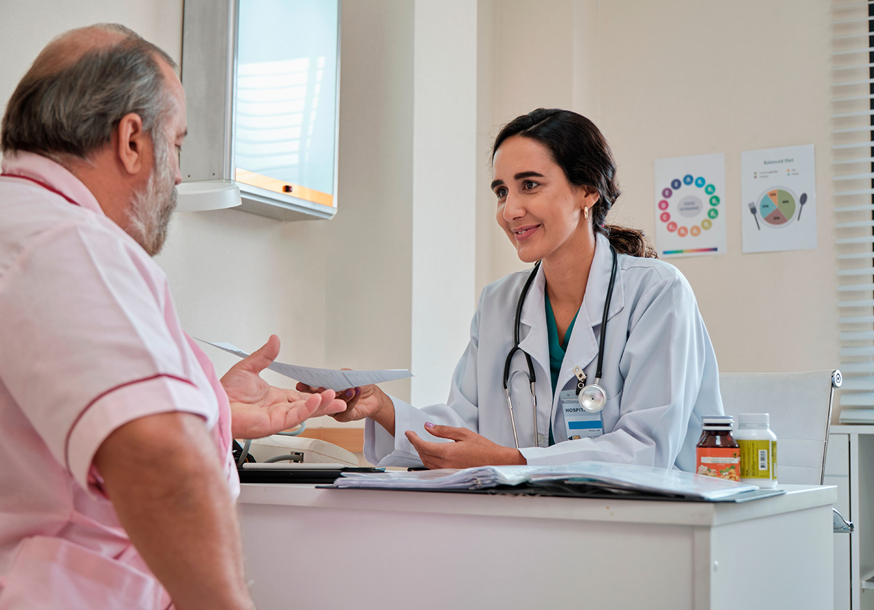 Médica atende sorridente um paciente em consulta. Ela usa estetoscópio e prontuários sobre a mesa do seu consultório de nutrologia.