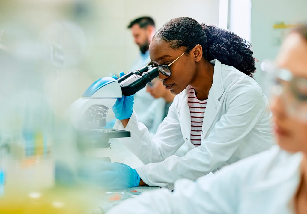 Ambiente de perícia médica com mulher analisando amostra no microscópio de um laboratório e outros profissionais ao redor.