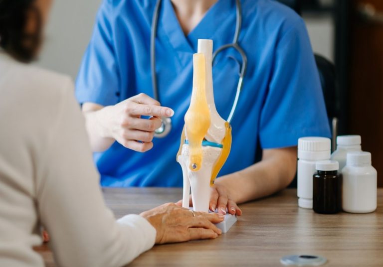 Médica de jaleco aponta para um modelo de joelho na frente de uma paciente. Curso: Prótese de Joelho Infectada.