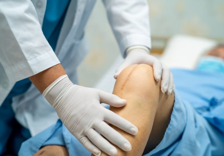 Um médico de jaleco e luvas brancas examina o joelho de um paciente em um ambiente hospitalar. Curso: Prótese Primária de Joelho.