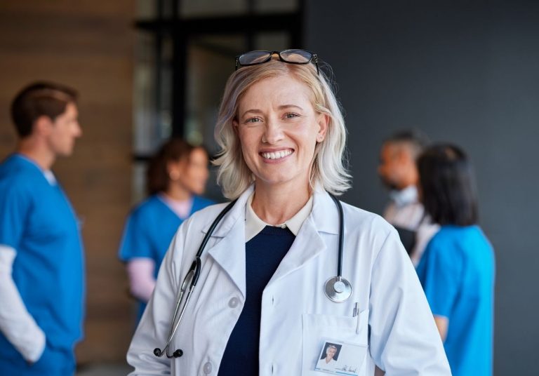 Médica sorridente com estetoscópio, outros profissionais ao fundo em ambiente hospitalar. Curso: Saúde Baseada em Valor.