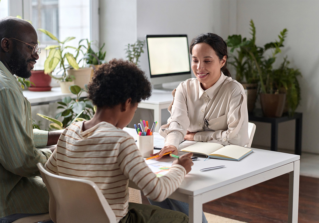 Família em consulta de saúde mental e atenção psicossocial, com criança desenhando e adultos conversando em um consultório com plantas e computador.