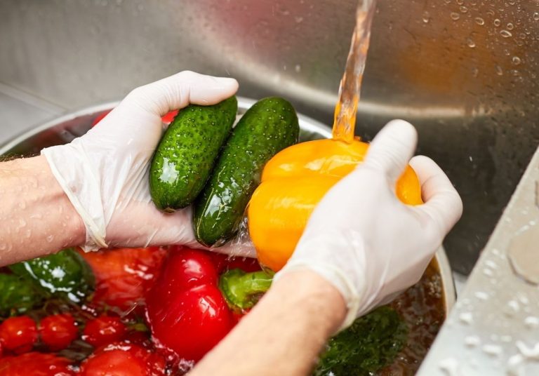 Mãos com luvas lavando pepinos e pimentões em pia com água. Curso: Segurança Alimentar e Manipulação de Alimentos.
