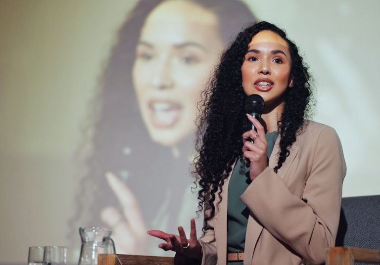 Mulher falando com microfone em apresentação, com sua imagem projetada em um telão ao fundo. Curso: Ser um Coach.