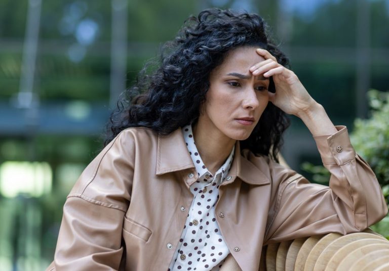Mulher com expressão preocupada, mão na testa, sentada em um banco ao ar livre. Curso: Superar o Medo no Ambiente de Trabalho.