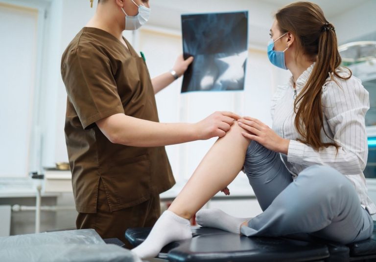 Médico mostrando raio-X do joelho a uma paciente, examinando seu joelho em consultório. Curso: Traumatologia do Joelho.