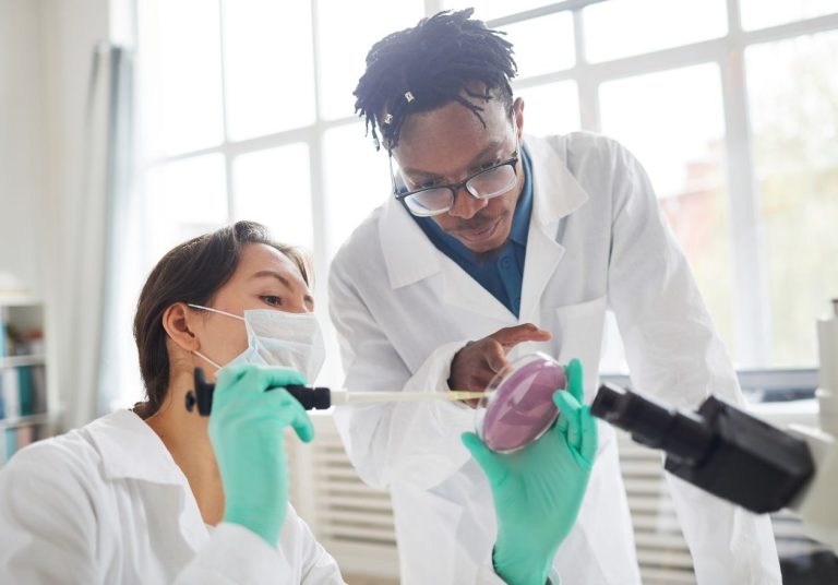 Dois profissionais em jalecos e luvas, analisando amostra em microscópio, em laboratório. Curso: Trilha de Aprendizagem: Infectologia.