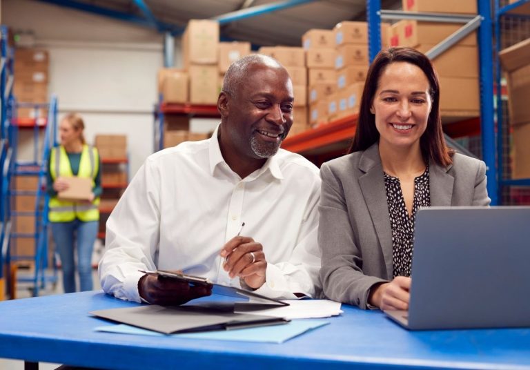 Duas pessoas sorrindo, trabalhando com laptop e documentos em um depósito com caixas ao fundo. Curso: Tudo Sobre o Supply Chain.