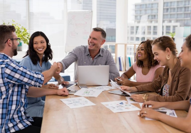 Equipe sorri e aperta as mãos em reunião com laptop e papéis sobre a mesa. Curso: Visão Compartilhada.