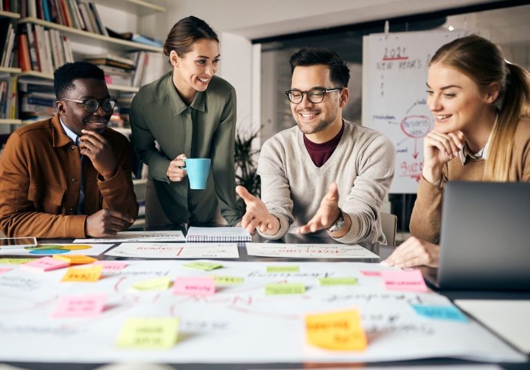 Equipe discute em mesa com papéis, post-its e laptop, em ambiente criativo, com estantes. Curso: Visão Sistêmica.