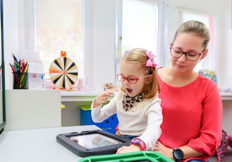 Mulher e criança com tablet em sala com brinquedos e materiais educativos. Curso: Capacitação em Comunicação Aumentativa e Alternativa.
