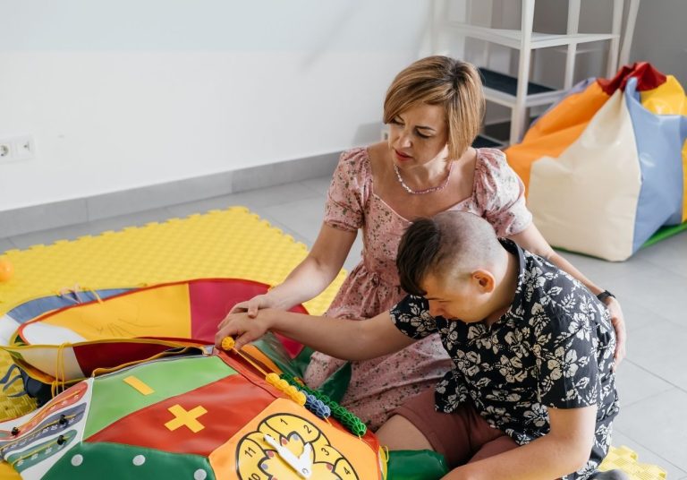 Pessoas brincam em tapete colorido com brinquedos sensoriais em sala com puffs. Curso: Capacitação em Integração Sensorial - Terapeuta Ocupacional.