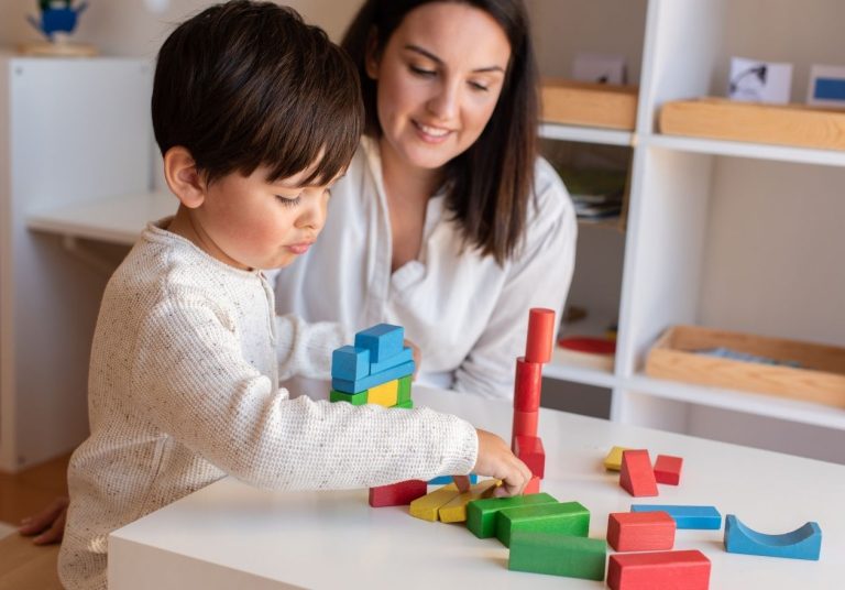 Mulher e criança brincam com blocos coloridos à mesa, em sala com estantes. Curso: Capacitação em Terapias Complementares.