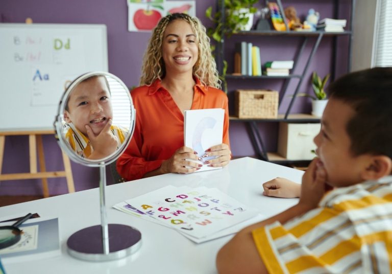 Mulher e criança em sala com espelho e cartões, quadro e estantes ao fundo. Curso: Capacitação em Transtornos Motores de Fala.