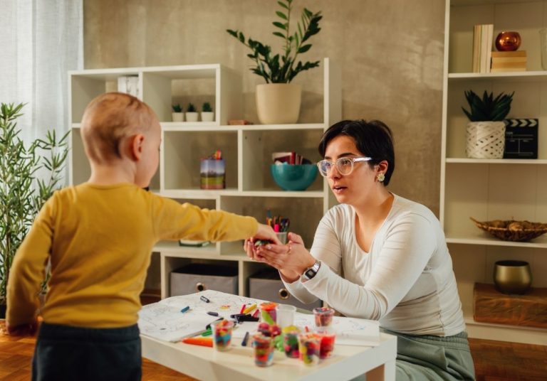 Mulher e criança interagem com objetos coloridos em mesa, em sala com plantas e estantes. Curso: Capacitação em VBMAPP e Programação Terapêutica.