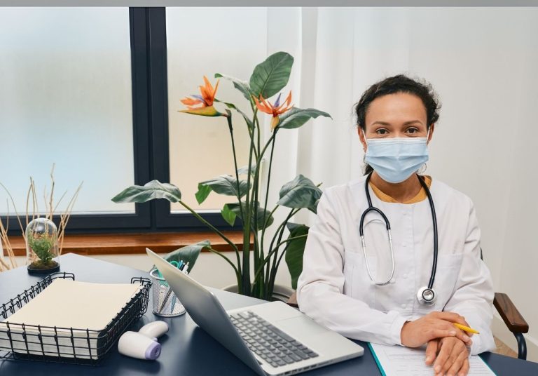 Médica com máscara em consultório com laptop, caderno e planta ao fundo. Curso: Capacitação Médicos Cooperados: Transtorno Globais do Desenvolvimento na Saúde Suplementar.