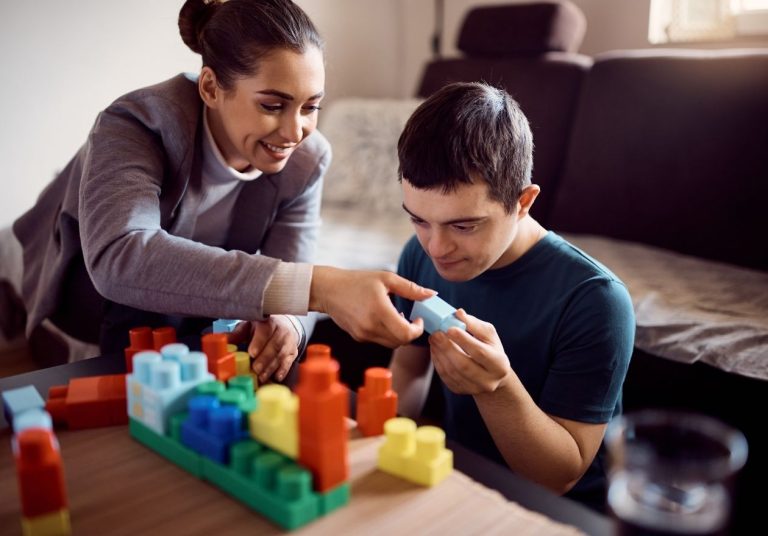 Mulher orienta garoto com blocos coloridos em mesa, em ambiente com sofá ao fundo. Curso: Capacitação para Aplicador ABA.