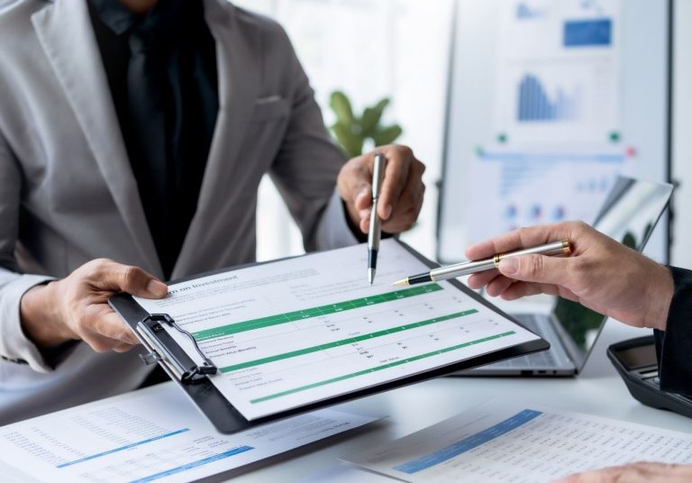 Pessoas analisam prancheta com dados financeiros em escritório com gráficos e planta. Curso: Impactos Financeiros e Atuarial dos Transtornos Globais do Desenvolvimento na Saúde Suplementar.
