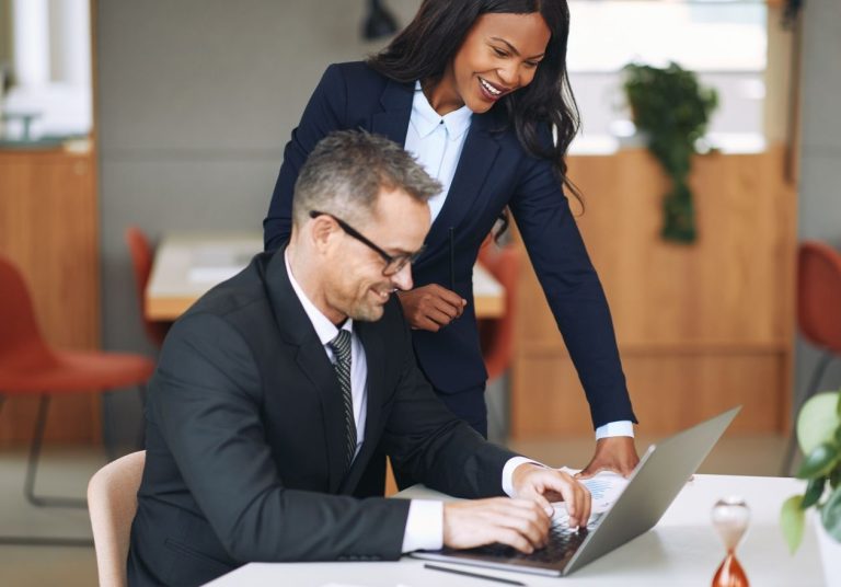 Duas pessoas em trajes formais analisam um laptop em uma mesa, em um ambiente profissional. Curso: Desenvolvimento para Dirigentes de Mercado em Cooperativas de Saúde.