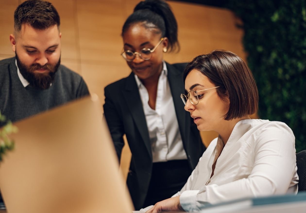 Três pessoas em um escritório, uma mulher sentada à mesa, um homem e uma mulher de pé. Curso: Formação Executiva em Governança Digital.