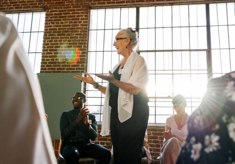 Uma mulher de pé gesticula e fala para um grupo sentado em uma sala com parede de tijolos. Curso: Gerenciar a Energia Emocional de uma Equipe.