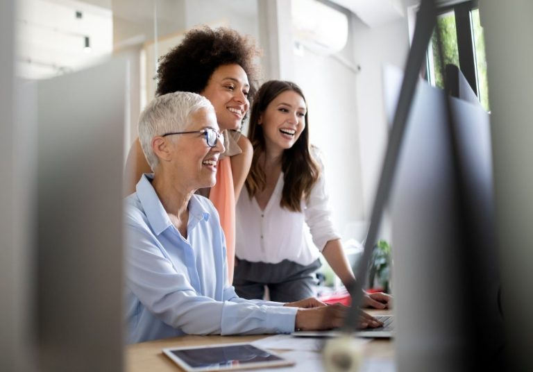 Três mulheres de diferentes gerações sorriem enquanto trabalham em um computador no escritório. Curso: Gerenciar Gerações.