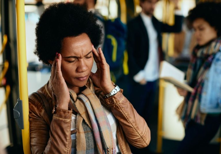 Uma mulher com os dedos nas têmporas parece muito estressada dentro de um ônibus lotado, com pessoas ao fundo. Curso: Gerenciar o Estresse.