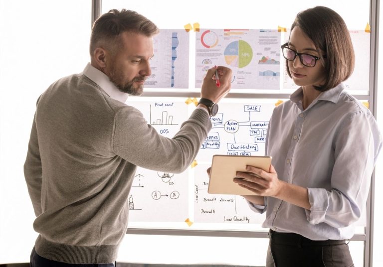 Homem e mulher conversam em frente a um quadro com gráficos e anotações. A mulher está segurando um tablet. Curso: Gestão Administrativa e Financeira.